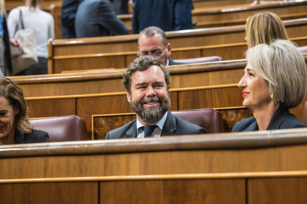 Gruppe von Menschen, die in einem Gerichtssaal sitzen, mit einem Mann in einem Anzug und zwei Frauen in legerer Kleidung, die nebeneinander sitzen, sowievisible Bänke im Hintergrund.