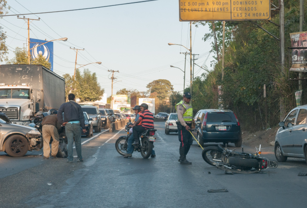 Gruppe von Menschen um ein verunglücktes Motorrad am Straßenrand mit mehreren Fahrzeugen, darunter ein Lastwagen, im Hintergrund und Bäumen, Masten, Lichtern, Schildern und Himmel.