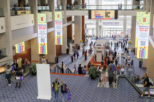 Eine große Menschenmenge, die durch ein großes Gebäude mit Bannern, Topfpflanzen, Rolltreppen, Geländern, Säulen, Texttafeln und Glastüren geht, wobei einige Taschen tragen, beim Nationalen Buchfestival.