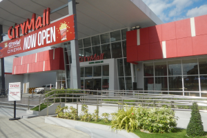 Eingang eines Stadtzentrums mit einem "Jetzt Geöffnet"-Schild, umgeben von Grün, einem Metallzaun, Menschen, einem Auto, Pfählen, einer Fahne, Lichtern und einem bewölkten Himmel.