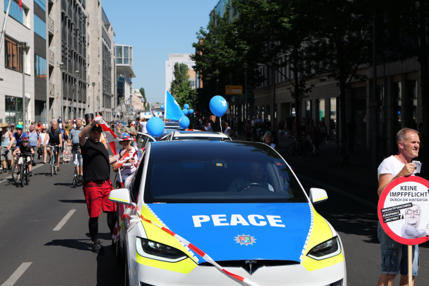 Eine Gruppe von Menschen, die auf Fahrrädern eine Straße entlangfahren, mit einem Polizeiwagen im Vordergrund, der Text anzeigt, und einer Person, die eine Tafel mit einem Bild und Text hält, vor einem Hintergrund aus Bäumen, Gebäuden mit Fenstern, Laternenpfählen und einem klaren blauen Himmel.
