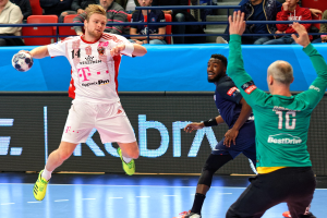 Eine Gruppe von Männern, die auf einem Feld Handball spielen, während im Hintergrund Zuschauer zu sehen sind.