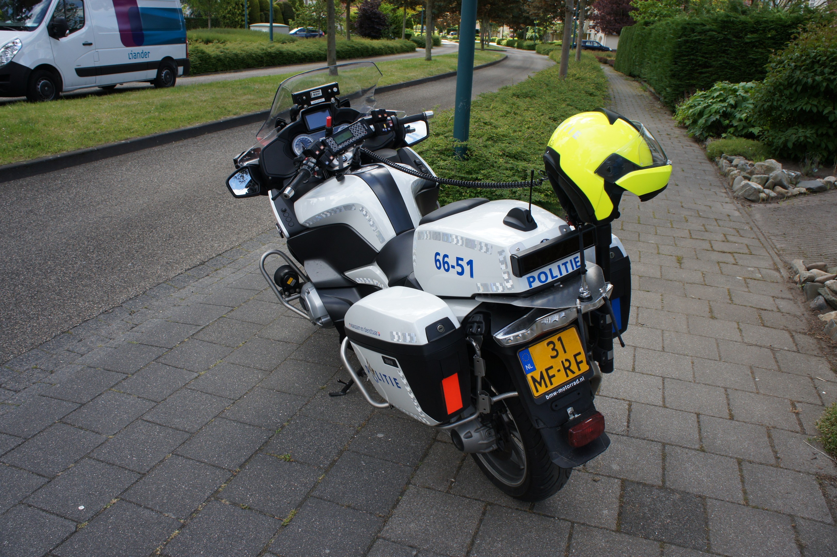 Ein auf der Seite der Straße geparktes Polizeimotorrad mit einem Helm darauf, umgeben von Bäumen, Pfählen, Gebäuden, Pflanzen, Gras, Steinen und anderen Fahrzeugen.
