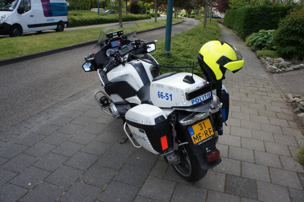 Ein auf der Seite der Straße geparktes Polizeimotorrad mit einem Helm darauf, umgeben von Bäumen, Pfählen, Gebäuden, Pflanzen, Gras, Steinen und anderen Fahrzeugen.