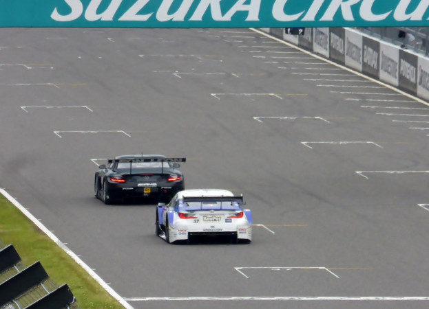 Zwei Sportwagen auf der Suzuka-Circuit-Rennstrecke, mit Gras und Metallobjekten auf der linken Seite und Tafeln mit Text auf der rechten Seite.
