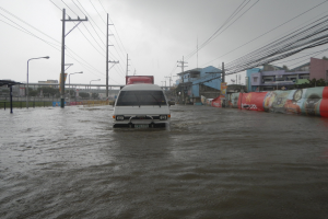 Ein Van fährt durch eine überflutete Stadtstraße mit Strommasten, Laternenmasten und Gebäuden auf beiden Seiten und einer Brücke im Hintergrund und dem Himmel darüber.