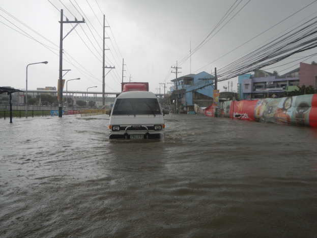 Ein Van fährt durch eine überflutete Stadtstraße mit Strommasten, Laternenmasten und Gebäuden auf beiden Seiten und einer Brücke im Hintergrund und dem Himmel darüber.