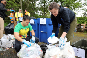 Eine Gruppe von Menschen in einem Park, die Müll sammeln, darunter ein Mann und eine Frau in der Mitte, die Handschuhe tragen und Teller halten, mit Plastikabdeckungen, Flaschen und anderem Müll auf dem Boden, einem Mülleimer und einer hölzernen Bank auf der rechten Seite und Bäumen und einem klaren blauen Himmel im Hintergrund.