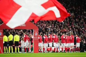 Gruppe von Menschen auf einem Fußballfeld mit einer roten und weißen Flagge im Vordergrund, einem Bogen mit "Bayern München vs. Bayern München Wetten & Vorschau" im Hintergrund und einer großen Menge im Stadion.