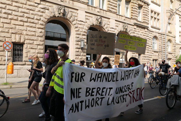 Eine Gruppe von Menschen marschiert bei einer Protestaktion gegen die Anti-Kriegs-Bewegung in Berlin, einige halten Schilder und andere fahren Fahrräder, mit einem Gebäude im Hintergrund, das Fenster, Bögen, Säulen und Skulpturen aufweist.