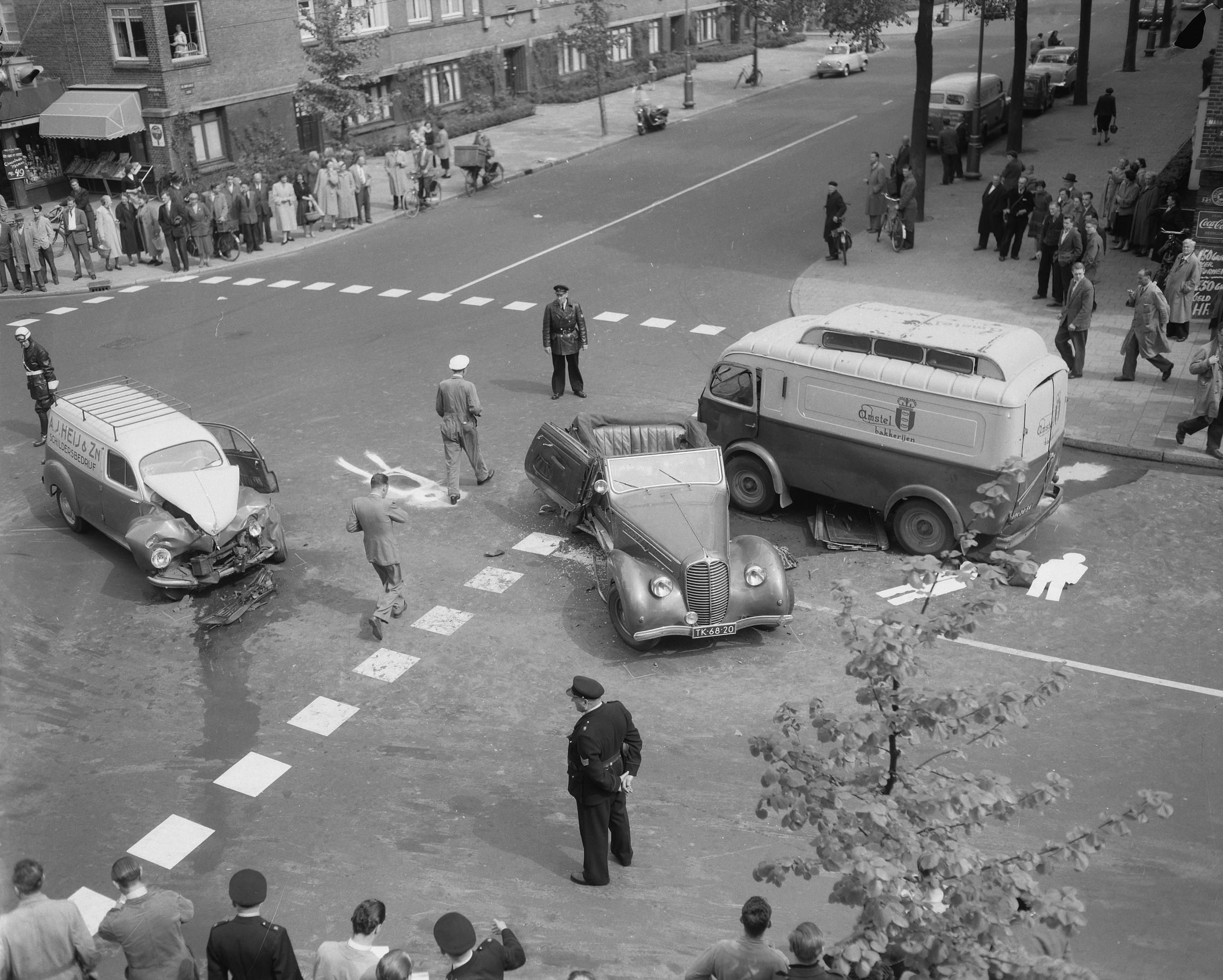 Schwarz-weiß-Szene eines Autounfalls an einer Straßenkreuzung mit einem Van, anderen Fahrzeugen und einer Gruppe von Schaulustigen, vor Häusern, Bäumen, Pfählen und Texttafeln.