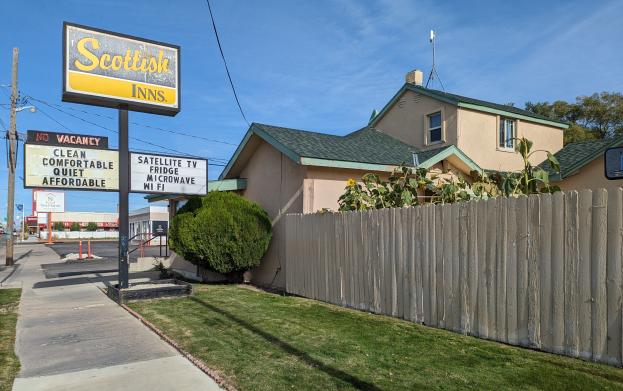 Ein schottisches Motel in San Francisco, Kalifornien, mit einem Haus mit Dach und Fenstern, einem hölzernen Zaun, Pflanzen, Gras, Bäumen, Schildern, Strommasten mit Drähten, einer Straßenlaterne, Fahrzeugen auf der Straße und einer bewölkten Himmel.