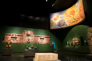 Ein Ausstellungsraum im Anthropologischen Museum von Mexiko-Stadt mit verschiedenen Artefakten, darunter Skulpturen, Tischgegenstände, eine Texttafel, eine Wandmalerei und Deckenleuchten.