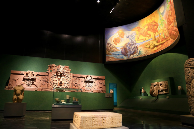 Ein Ausstellungsraum im Anthropologischen Museum von Mexiko-Stadt mit verschiedenen Artefakten, darunter Skulpturen, Tischgegenstände, eine Texttafel, eine Wandmalerei und Deckenleuchten.