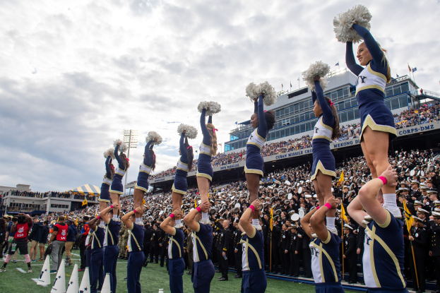 Eine Gruppe von Cheerleadern in blauen und weißen Uniformen führt einen Stunt aufeinander aus, während sie Pompons halten, mit einer großen Menge und einem Stadion im Hintergrund.