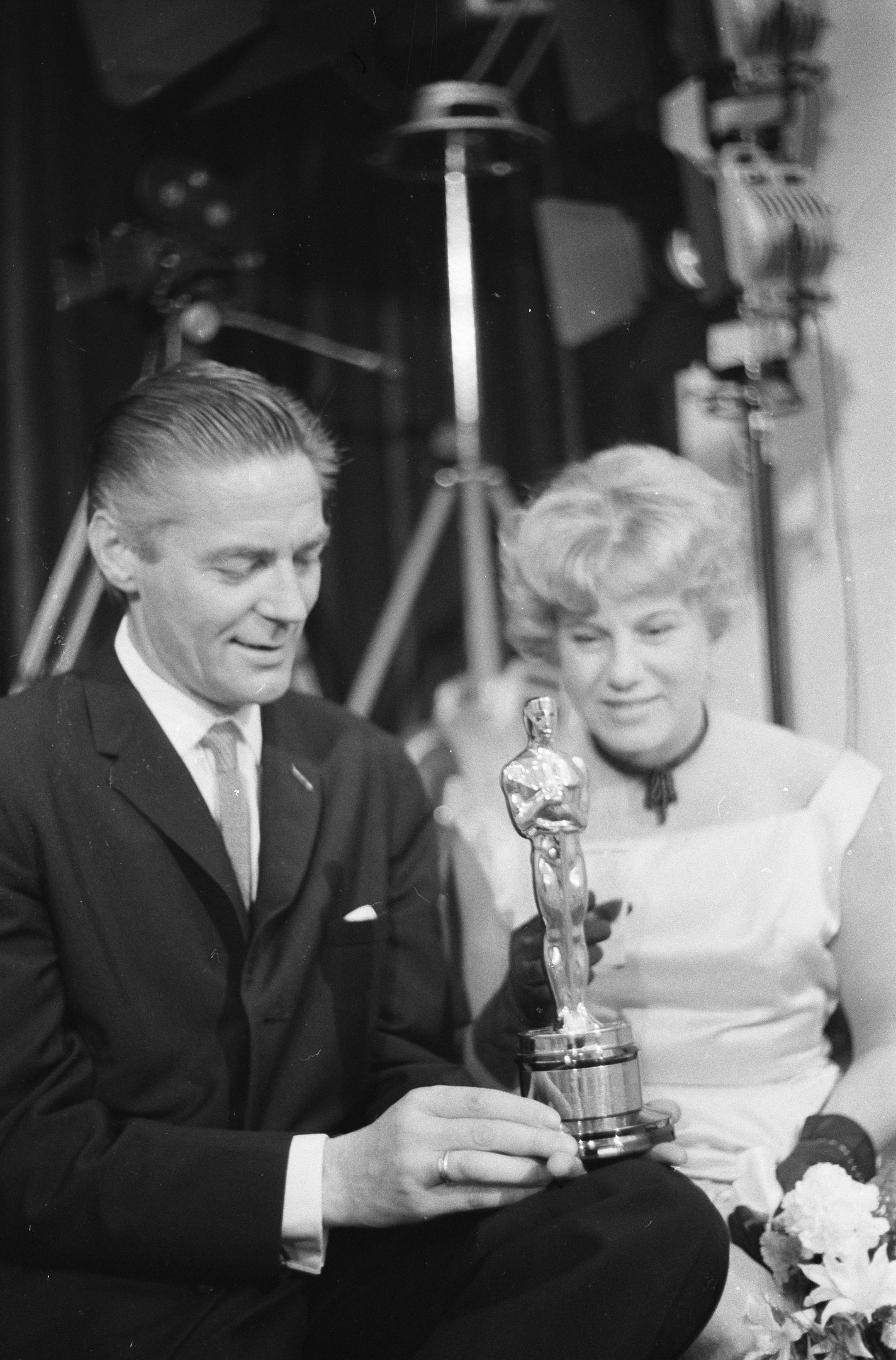 Ein Schwarz-Wei├č-Bild eines formell gekleideten Mannes mit einer Oscar-Statue und einer Frau mit einem Blumenstrau├čus, die nebeneinander sitzen, mit beleuchteter Kulisse im Hintergrund.