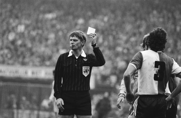 Schwarzes und weißes Bild eines Schiedsrichters mit roter Karte, mit einer Menge Menschen in Fußballtrikots hinter einem Zaun.