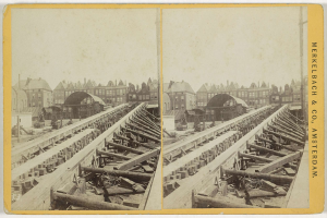 Schwarz-weiß-Fotografie einer Bahntrasse mit Gebäuden im Hintergrund, mit der Aufschrift "Merkelbach & Co, Co. Amsterdam" auf der rechten Seite und verstreuten Holzgegenständen im Vordergrund.
