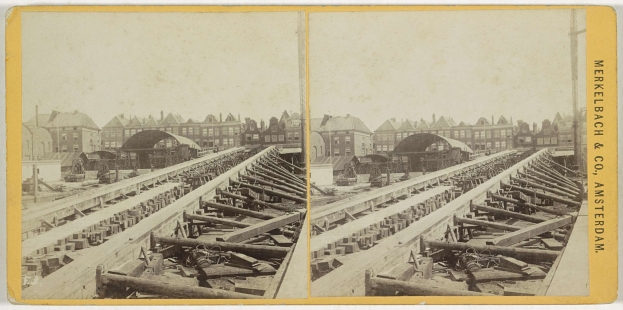 Schwarz-weiß-Fotografie einer Bahntrasse mit Gebäuden im Hintergrund, mit der Aufschrift "Merkelbach & Co, Co. Amsterdam" auf der rechten Seite und verstreuten Holzgegenständen im Vordergrund.