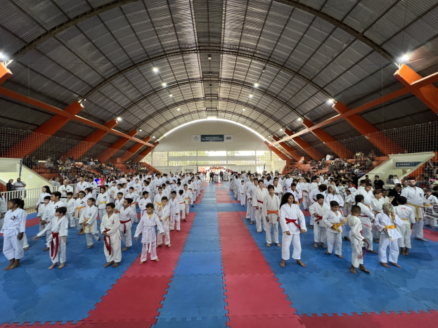 Große Gruppe von Menschen auf einer blauen und roten Matte in einer Turnhalle mit Zuschauern auf der Treppe und Deckenlampen, Hintergrundtafeln wahrscheinlich für ein Taekwondo-Event.