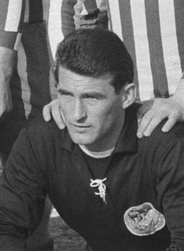 Schwarzes und weißes Foto eines Mannes, der auf einem grünen Fussballfeld sitzt und ein T-Shirt und eine Kette trägt, mit zwei Personen dahinter.
