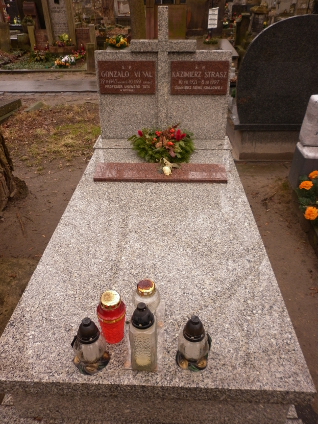 Ein Grabstein auf einem Friedhof, geschmückt mit einem Blumenstrauß und mehreren Gläsern, umgeben von anderen Gräbern, Bäumen und Blumen.