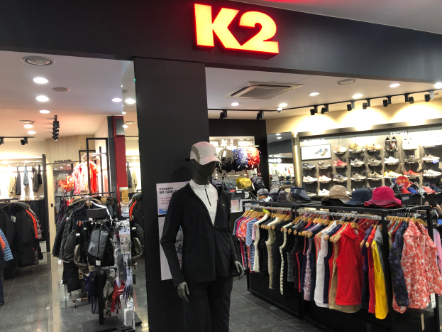 Ein K2-Kleidungsgeschäft in Singapur mit Schaufensterpuppen in Mützen, Kleidung an Bügeln, Schuhe auf Gestellen und einer Wandtafel mit Text.