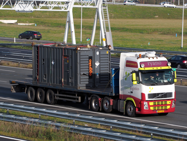 Ein Lkw mit einem große Behälter fährt auf einer Autobahn, mit einigen anderen Fahrzeugen, Masten, Bäumen, Gebäuden und einem klaren blauen Himmel im Hintergrund.