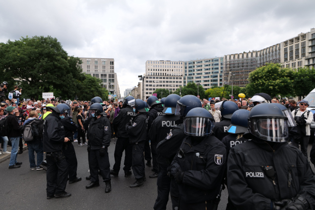 Große Gruppe von Polizisten vor einer Menschenmenge während einer Demonstration in Berlin, Deutschland, mit Kameras in der Menge und Bäumen, Gebäuden und einem bewölkten Himmel im Hintergrund.