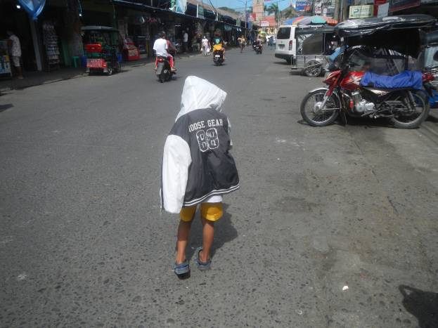 Ein junger Junge geht eine Straße voller Motorräder und anderer Fahrzeuge entlang, flankiert von Gebäuden mit Schildern, unter einem klaren blauen Himmel mit Bäumen und Pfosten im Hintergrund.
