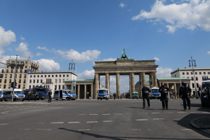 Eine Gruppe von Polizeibeamten steht vor dem Brandenburger Tor in Berlin, Deutschland, das mit Säulen, einer Statue und umgeben von Gebäuden mit Fenstern, Fahrzeugen, Laternenmasten und Verkehrszeichen unter einem bewölkten Himmel geschmückt ist.