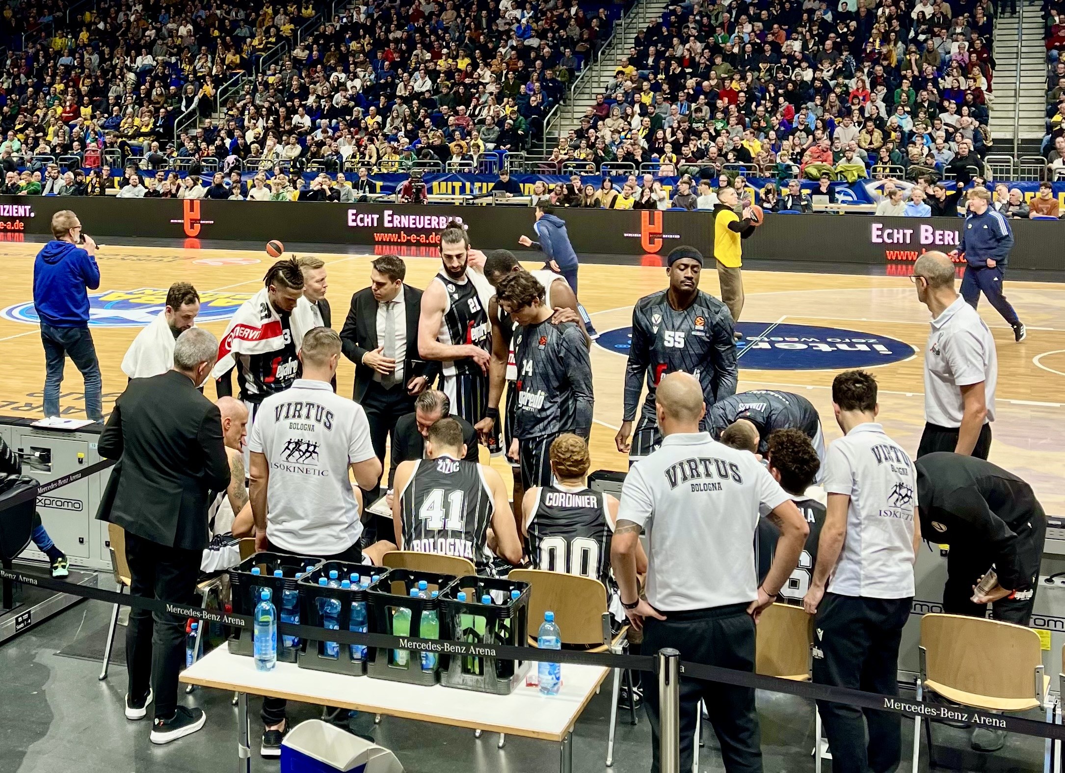 Eine Gruppe von Männern auf einem Basketballfeld mit einer Menge, die auf Stühlen und Stehplätzen sitzt und steht, ein Fotograf mit Equipment auf einem Tisch links und Hintergrundtafeln mit den Teamnamen Virtus Berlin und Echt Berlin.