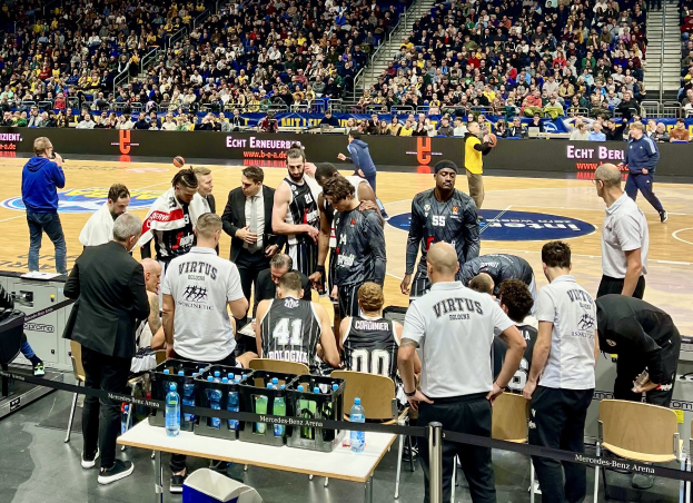 Eine Gruppe von Männern auf einem Basketballfeld mit einer Menge, die auf Stühlen und Stehplätzen sitzt und steht, ein Fotograf mit Equipment auf einem Tisch links und Hintergrundtafeln mit den Teamnamen Virtus Berlin und Echt Berlin.