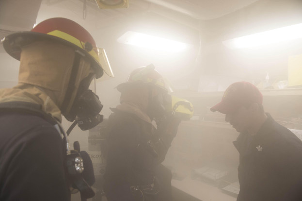 Feuerwehrleute in Helmen und Ausrüstung atmen Rauch während einer Trainingssitzung aus, wobei einer eine Kamera hält und Hintergrundlichter zu sehen sind.