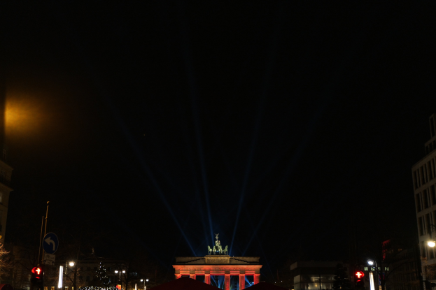 Das Brandenburger Tor in Berlin nachts, beleuchtet von Straßenlaternen, mit Gebäuden, Bäumen, Pfählen, Verkehrszeichen, Schildern und Zelten drumherum vor einem dunklen Himmel.