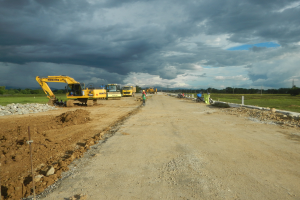 Ein Schotterweg mit Baumaschinen wie Kr├Ąne, Steine, Masten und Menschen, mit B├Ąumen, Gras und einem bew├Âlktem Himmel im Hintergrund.