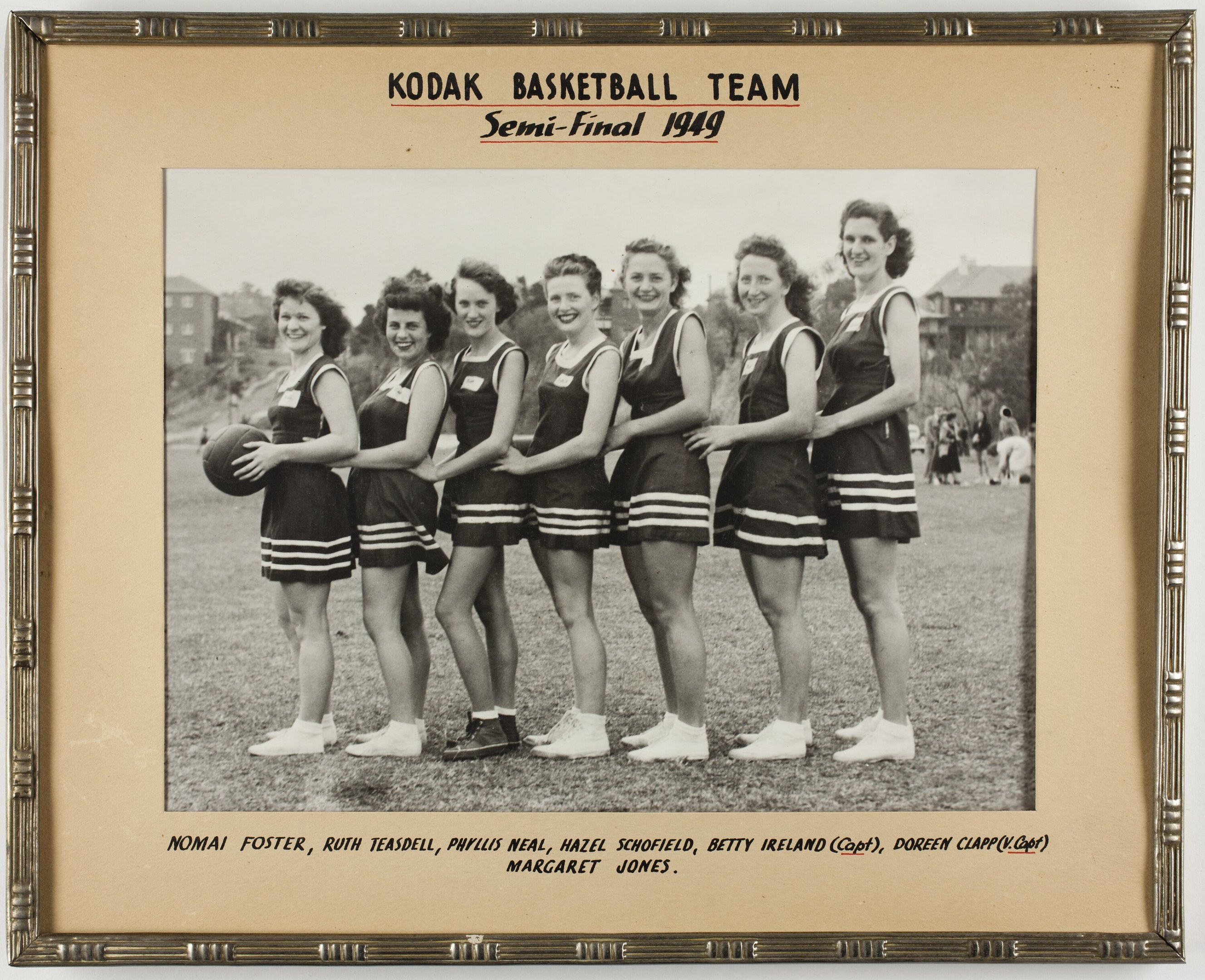Schwarzes und weißes Foto einer Gruppe von Frauen, die lächeln und einen Basketball halten, mit Menschen, Bäumen, Gebäuden und Himmel im Hintergrund, beschriftet mit "Kodak Basketball Team Semi-Final 1949".