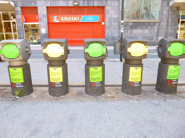 Reihe von Parkautomaten entlang einer Straße mit einem Gebäude mit Glasfenstern und einer Rolllade mit Text im Hintergrund und einem nahen Müllcontainer.