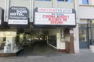 Das Lincoln Theater in Berlin, Deutschland, ein Gebäude mit Glasfenstern und -türen und einer Tafel mit Text darauf, zeigt verschiedene Objekte im Inneren und vermittelt den Eindruck einer pulsierenden Stadtlandschaft.
