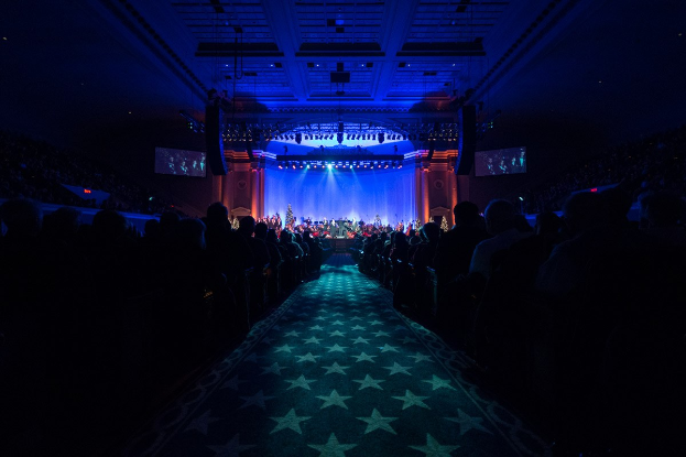 Ein großer Auditorium mit Menschen in Reihen vor einer Bühne, auf der Musiker spielen, unter einer Decke mit Lichtern und Fernsehern, was auf eine Konzertsäleinrichtung hinweist.