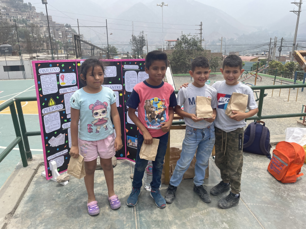 Gruppe von Kindern auf einem Basketballfeld mit Papiertüten in den H├Ąnden, mit Vorr├Ąten auf dem Boden, einer Tafel mit Text links daneben und einem eingez├Ąunten ├Ąu├čeren Bereich mit Geb├Ąuden und H├Ąngen im Hintergrund.