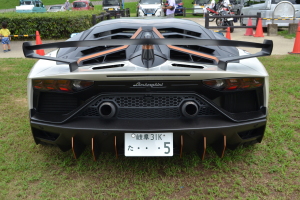 Ein Lamborghini Aventador LP700-4 Superveloce auf dem Goodwood Festival of Speed, mit einem Auto im Vordergrund, umgeben von Gras und Verkehrskegeln, Menschen auf der Straße links und Pflanzen, Bäumen, Gebäuden und einem klaren blauen Himmel im Hintergrund.