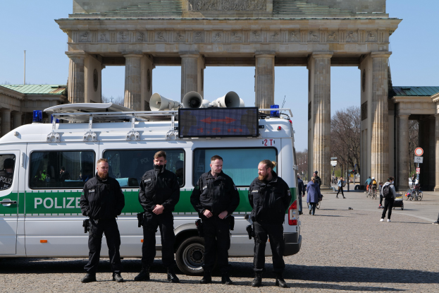 Polizisten in schwarzen Uniformen stehen auf einer Straße vor dem Brandenburger Tor in Berlin, mit einem weißen und grünen Fahrzeug im Vordergrund und Fußgängern und Radfahrern im Hintergrund.