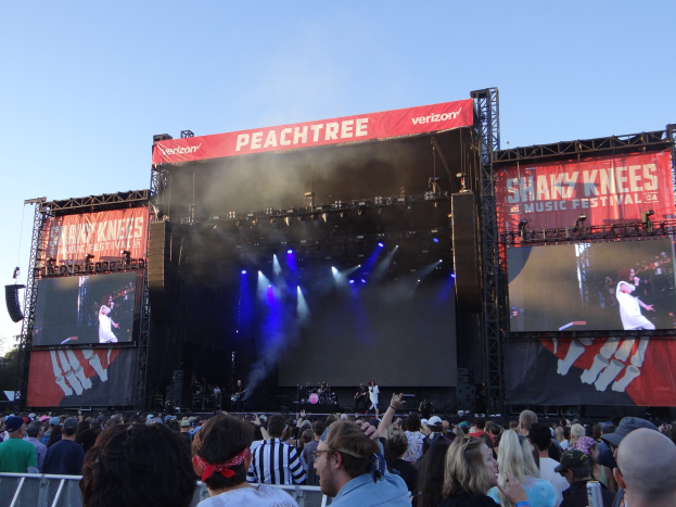 Eine große Menschenmenge steht vor einer hell erleuchteten Bühne auf dem Shaky Knees Music Festival, auf der Musiker unter Schildern mit Text spielen, mit einem Geländer im Vordergrund und einem klaren blauen Himmel im Hintergrund.