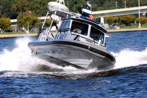 Ein Polizeiboot fährt über einen Gewässser mit einer Person darin, mit einer Brücke, Bäumen, Polen, Fahrzeugen, Gras und einem klaren blauen Himmel im Hintergrund.