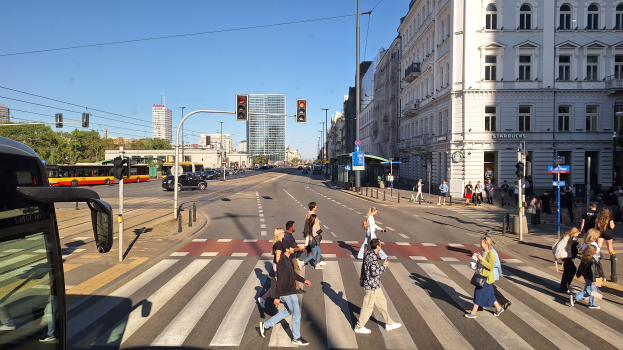 Eine Gruppe von Menschen, die die Straße an einer Ampel überqueren, mit einem Bus auf der linken Seite, Fahrzeugen auf der Straße, Verkehrszeichen, Laternenmasten, Gebäuden, Bäumen und einem klaren blauen Himmel.