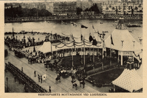 Ein altes Schwarz-Weiß-Foto einer Menge, die sich um einen Karneval mit Zelten, Fahnen, Stangen und Pferden im Vordergrund und Gebäuden, Bäumen und Wasser im Hintergrund versammelt hat, mit Text am unteren Bildrand.