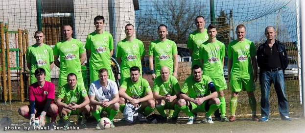Eine Gruppe von Männern in grünen T-Shirts steht auf einem Fußballfeld, einige hocken, mit einem Ball vor ihnen und einem Torpfosten rechts, vor einem Hintergrund aus Bäumen, Gebäuden und Fahrzeugen.