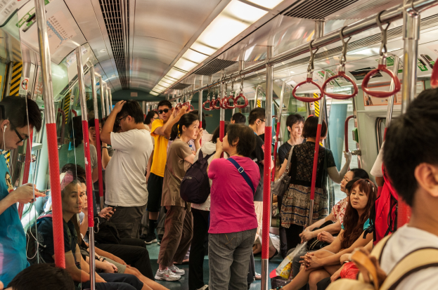 Menschen, die auf einem Zug fahren, einige sitzen auf Sitzen und andere stehen, mit Metallstangen, Griffen, Lampen und Plakaten im Inneren.