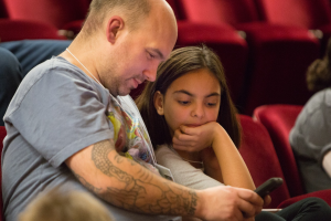 Ein Mann und ein junges Mädchen in einem Kinosaal, beide konzentriert auf ein Handy, mit anderen Sitzenden im Hintergrund und unscharfem Hintergrund.
